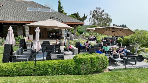 A patio with umbrellas and seating, where people relax among greenery and flowers near a house.