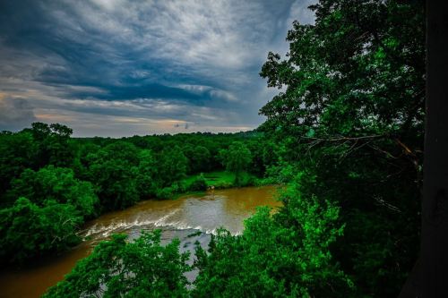 A serene river flows through lush green trees under a cloudy sky, creating a tranquil natural scene.