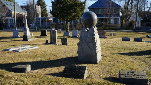 A cemetery scene featuring a prominent stone monument topped with a sphere, surrounded by various gravestones.