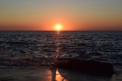 Sunset over the ocean, with waves gently splashing against a rocky shore, casting a warm glow on the water.