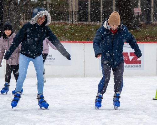 Two people skate on an ice rink in the snow, smiling and enjoying the winter activity.