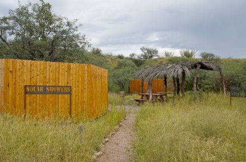 A pathway leads to solar showers, surrounded by tall grass and trees, with a shaded picnic area nearby.