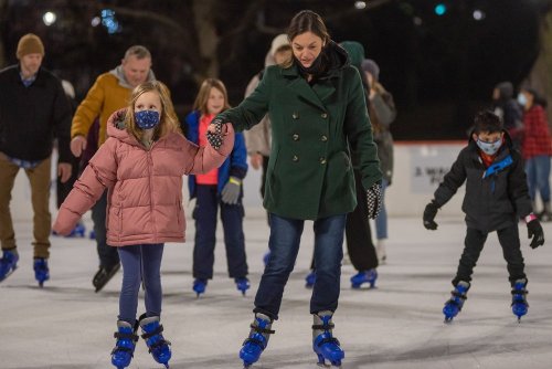 A woman and a girl skate together on an ice rink, surrounded by other skaters, all wearing masks.