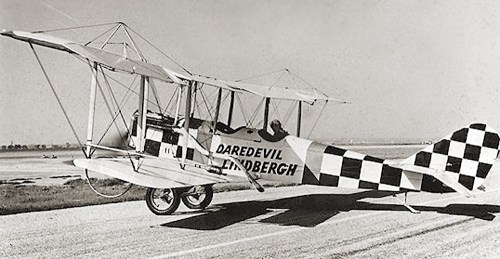 A vintage biplane with a checkered pattern, labeled "Daredevil Lindbergh," on a runway.