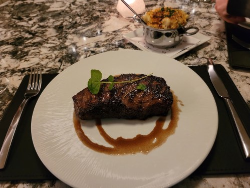 A perfectly cooked steak on a white plate, garnished with herbs, accompanied by a side dish in a metal bowl.