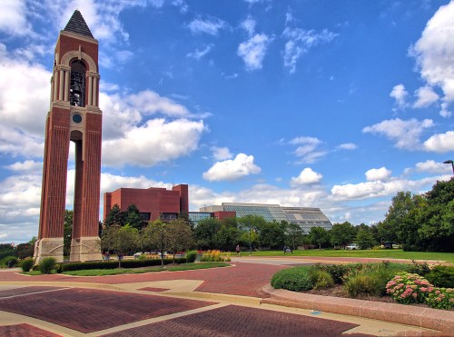 A brick clock tower stands in a landscaped area with a modern building and blue sky filled with clouds in the background.