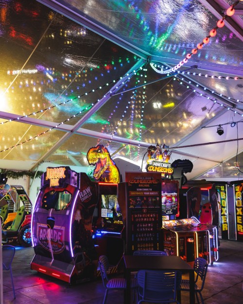 Colorful arcade games under a tent, featuring Jurassic Park and Minecraft, with festive lights reflecting above.