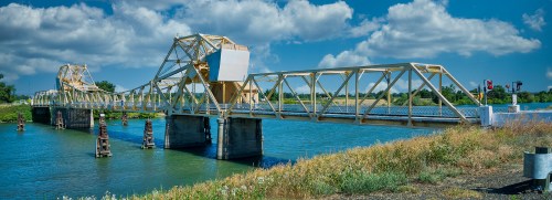 A yellow truss bridge spans a blue river under a partly cloudy sky, surrounded by green grass and trees.