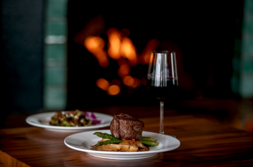 A gourmet meal featuring steak and asparagus, with a glass of red wine, set against a cozy fireplace backdrop.