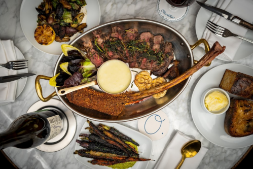 A beautifully arranged feast featuring grilled meats, vegetables, and sauces on a marble table with wine and utensils.