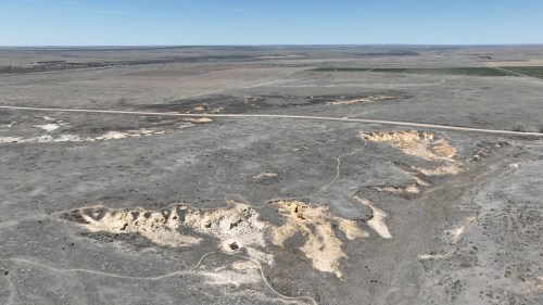 Aerial view of a vast, arid landscape with dry terrain and a winding path, surrounded by flat fields in the distance.