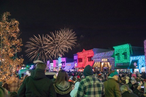 Crowd watching colorful fireworks over a festive, illuminated street decorated for the holidays at night.