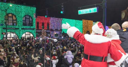 A festive crowd gathers under colorful holiday lights, with Santa waving from a stage.