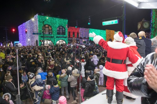 A crowd gathers at night, illuminated by colorful holiday lights, as Santa waves from a stage.