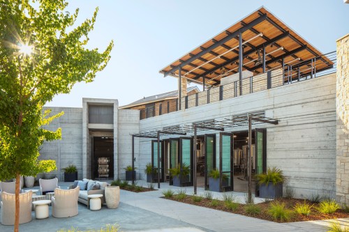 Modern building exterior with a rooftop terrace, surrounded by greenery and outdoor seating. Bright, sunny day.