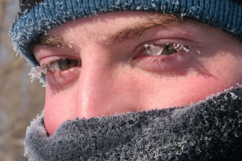 Close-up of a person’s face with frost on their eyelashes and a scarf, set against a cold, wintry background.