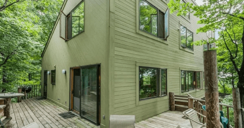 A modern green house surrounded by trees, featuring large windows and a wooden deck.