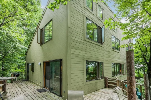 A modern green house surrounded by trees, featuring large windows and a wooden deck.