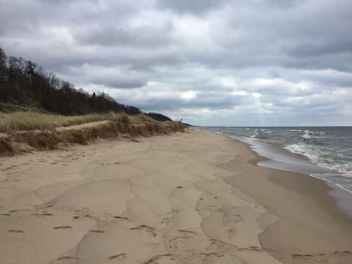 A sandy beach with gentle waves, grassy dunes, and overcast skies in the background.