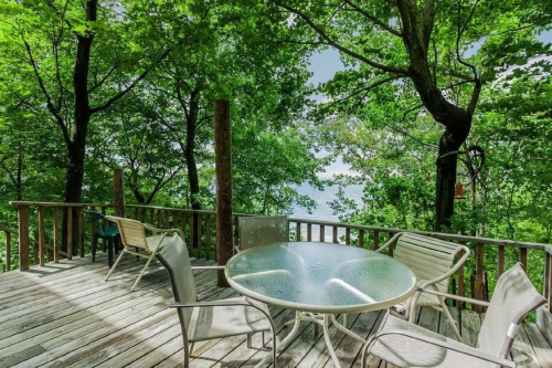 A wooden deck surrounded by trees, featuring a glass table and chairs, overlooking a serene body of water.