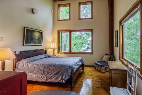 Cozy bedroom with a large bed, wooden floors, and windows overlooking greenery, featuring a lamp and a small chair.
