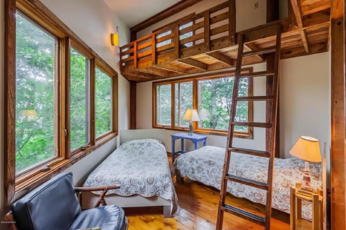 Cozy bedroom with two beds, wooden accents, a loft, and large windows overlooking greenery.