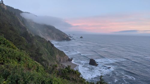 A scenic coastal view with cliffs, waves crashing on rocks, and a pastel sky at sunset.