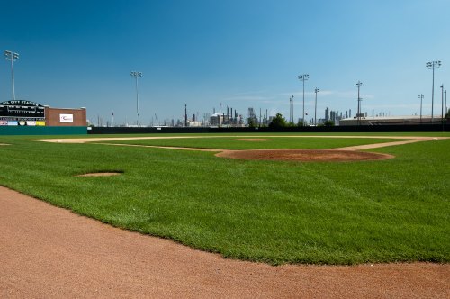 A baseball field with a well-maintained infield and outfield, set against a clear blue sky and industrial background.