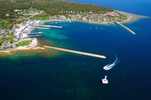 Aerial view of a coastal town with a marina, boats, and a lighthouse, surrounded by clear blue water and green hills.