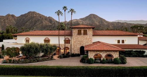 A large, elegant stone and stucco house with a red tile roof, set against a backdrop of mountains and palm trees.