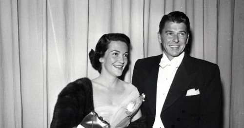 A smiling couple in formal attire, with the woman holding a bouquet, standing in front of a curtain backdrop.