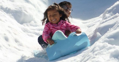 Two children joyfully sledding down a snowy hill, with one girl smiling widely in front and a boy behind her.