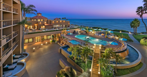 A coastal resort at sunset, featuring a pool, palm trees, and ocean views, with colorful umbrellas around the pool area.