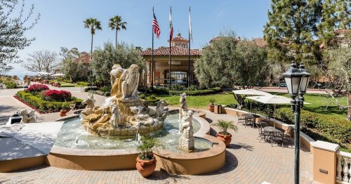 A scenic view of a resort entrance featuring a fountain, palm trees, and outdoor seating areas.