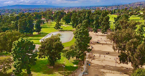 Aerial view of a park with green trees, a pond, and walking paths, bustling with people enjoying the outdoors.