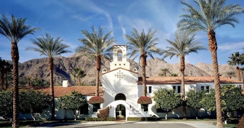A picturesque building surrounded by palm trees and mountains, featuring a bell tower and lush greenery in the foreground.