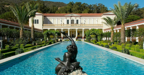A serene courtyard with a blue pool, palm trees, and a statue, framed by lush greenery and classical architecture.