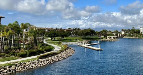 A serene waterfront view featuring a landscaped park, palm trees, and a calm blue lake under a partly cloudy sky.