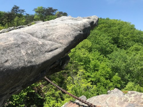 A rocky outcrop juts out over a lush green forest, with a chain attached for safety.