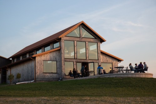 A modern wooden house with large windows, people gathered outside on a patio during sunset.