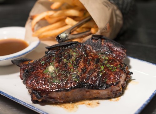 A grilled steak with a charred crust, garnished with herbs, served on a plate alongside a side of fries and dipping sauce.