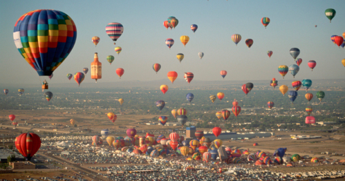 A vibrant sky filled with colorful hot air balloons over a crowded field and distant landscape.