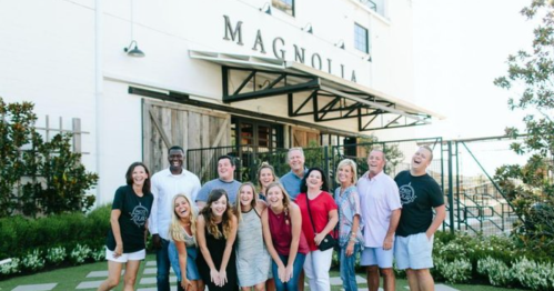 A group of 15 people smiling and posing together outside the Magnolia building, surrounded by greenery.
