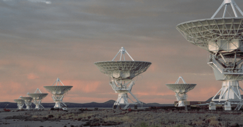 A row of large satellite dishes against a colorful sunset sky, set in a desert landscape.