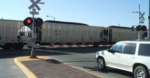 A white SUV stops at a railroad crossing as a freight train passes by, with red lights flashing.