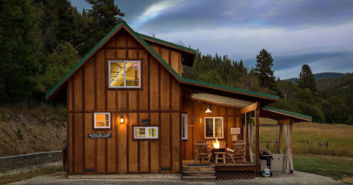A cozy wooden cabin surrounded by trees and mountains, with a porch and warm lights glowing from the windows.