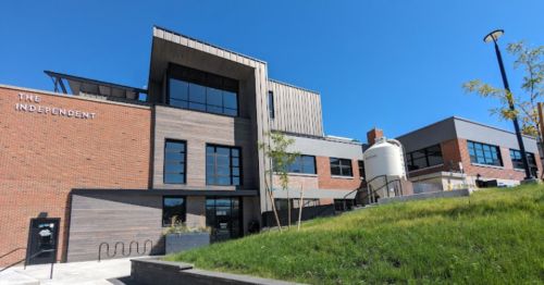 Modern building with a mix of brick and wood, featuring large windows and a grassy slope in front under a clear blue sky.