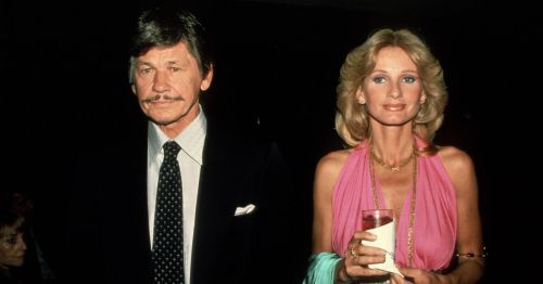 A man in a suit and a woman in a pink dress hold drinks, posing together at an event.