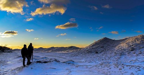 Two hikers silhouetted against a colorful sunset over a snowy landscape with rolling hills.
