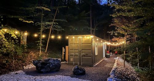 A cozy container cabin surrounded by trees, illuminated by string lights at dusk, with a gravel path and rocks.